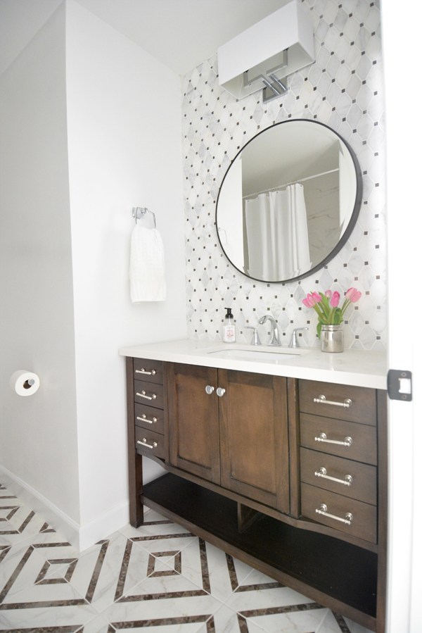There are pink tulips on the counter in this bathroom with wooden cabinets, a round mirror and a geometric patterned floor.
