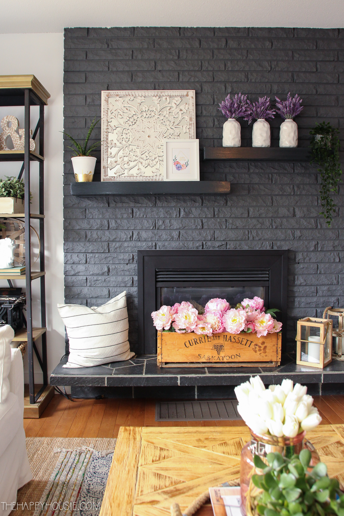 There are 3 white vases with lilac and a small wooden box with peonies on the fireplace mantel.