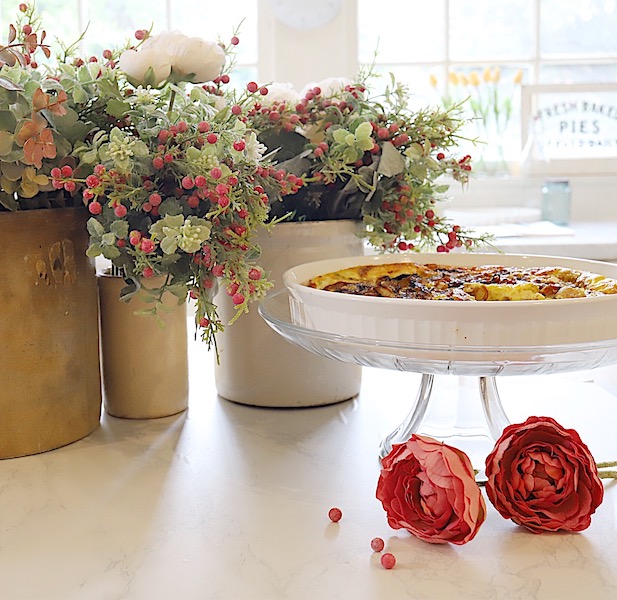 A quiche in a pie plate on top of a cake stand beside flowers on the counter.