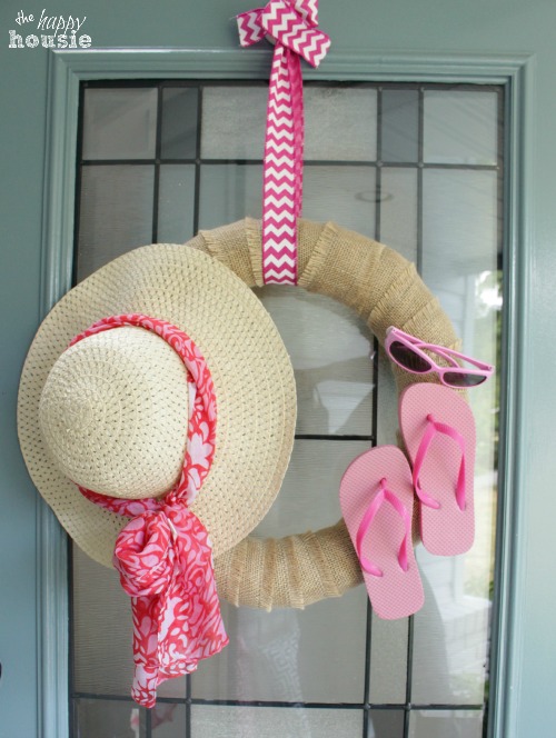 A wreath with a hat, flip flops and pink sunglasses hanging on a door.