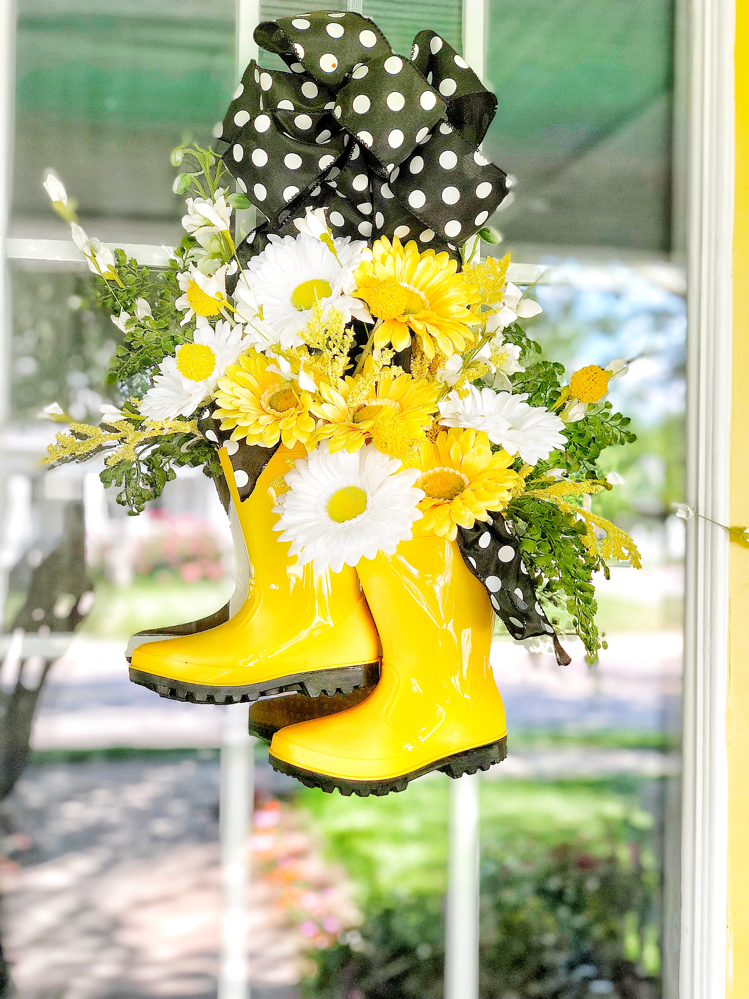 Yellow rain boots with flowers in it hanging in front of a window.