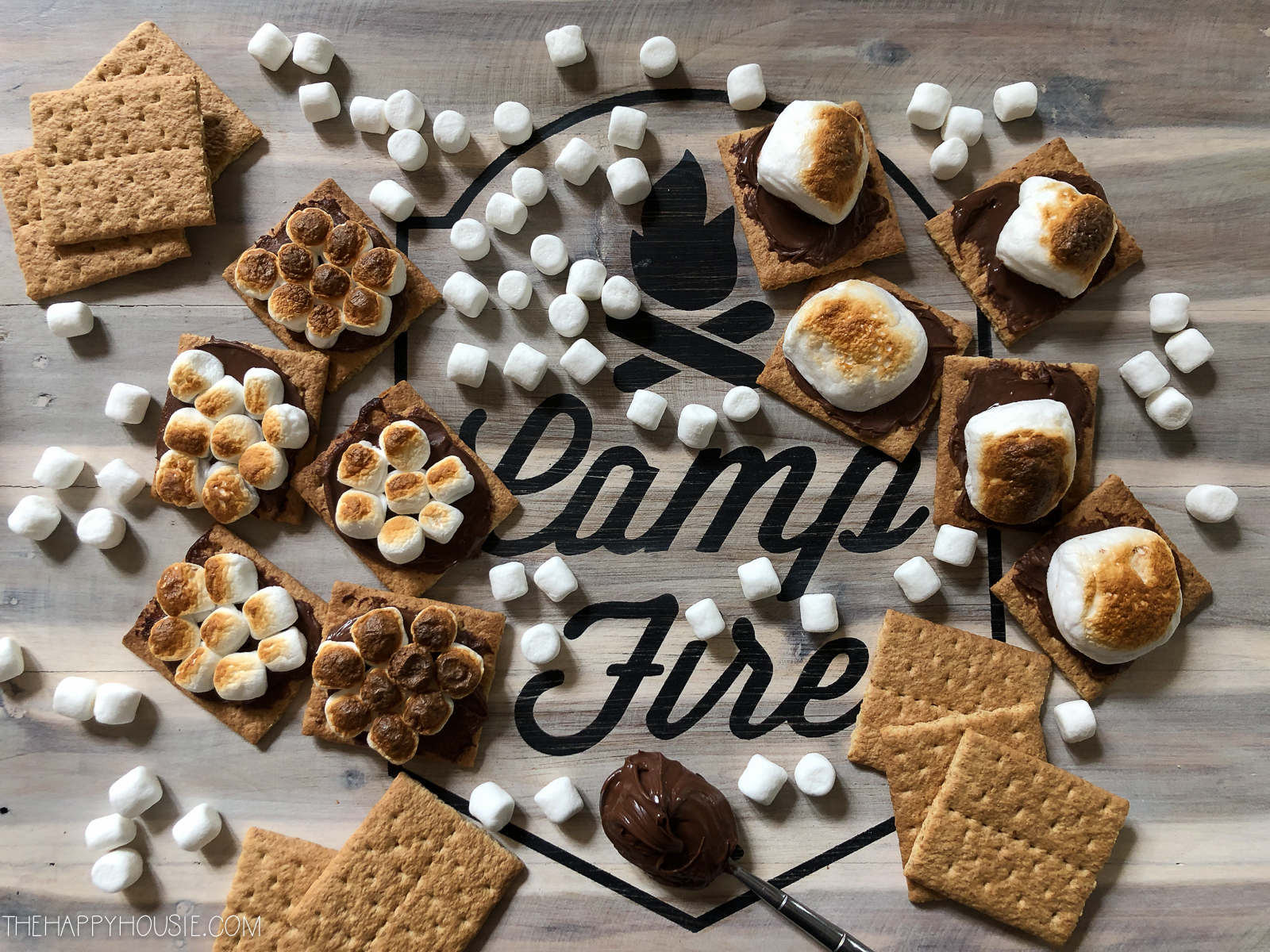 A wooden board that says camp fire on it with graham cookies, marshmallows and Nutella scattered around it.