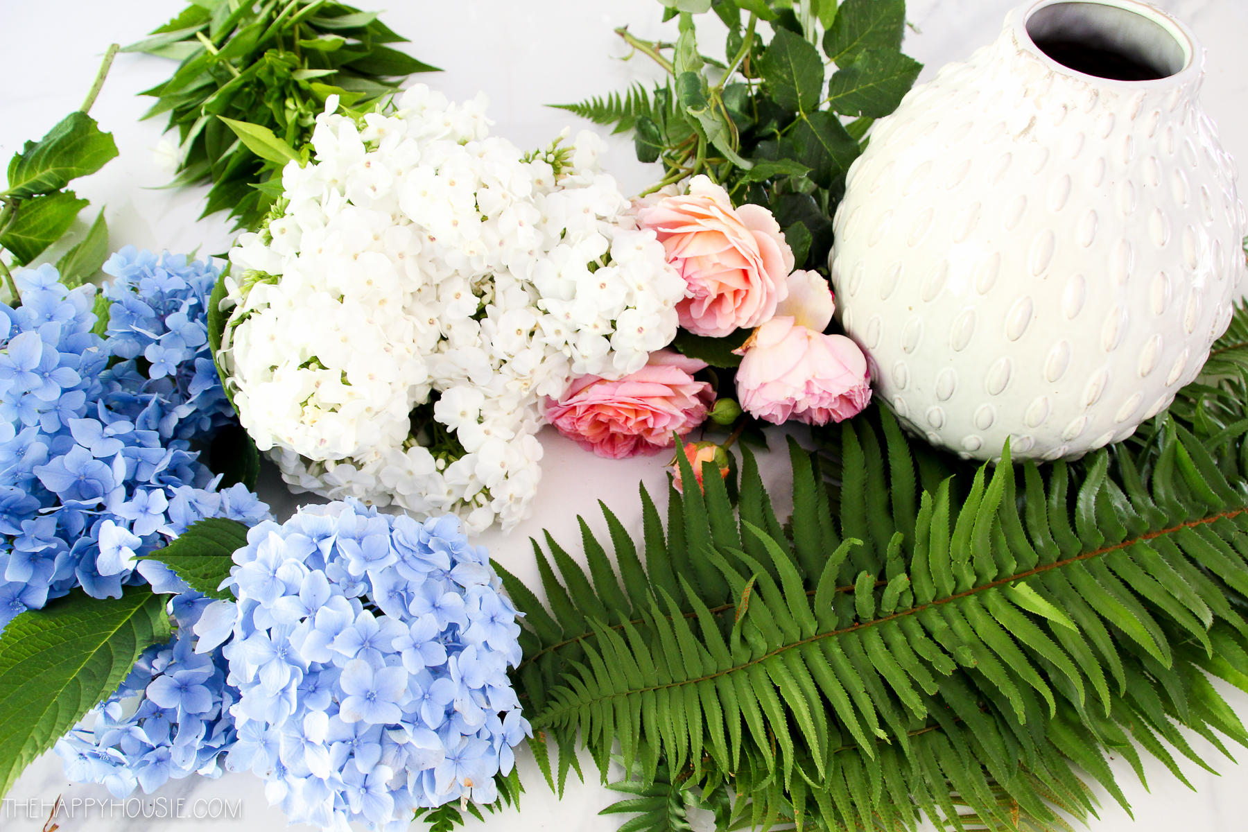 Pretty flowers and ferns laid out beside a white vase.