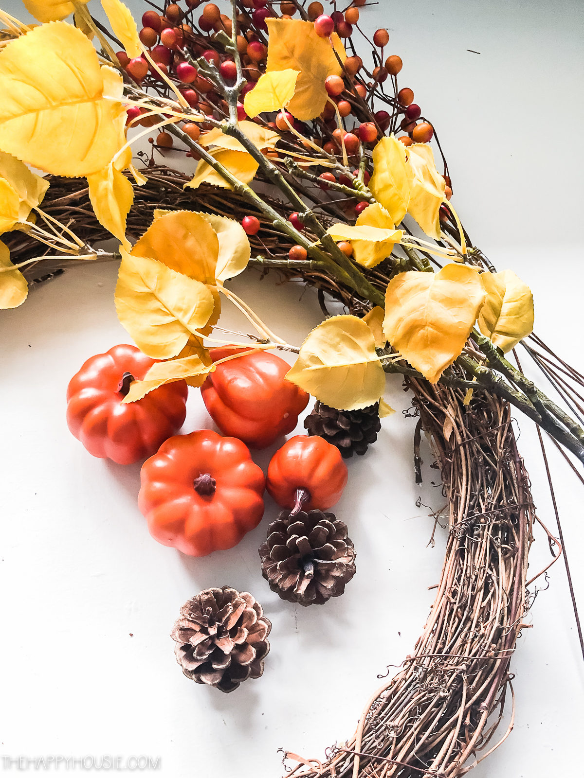 A grapevine wreath, yellow leaves, berries, pine cones, and mini pumpkins laid out to put on the wreath.