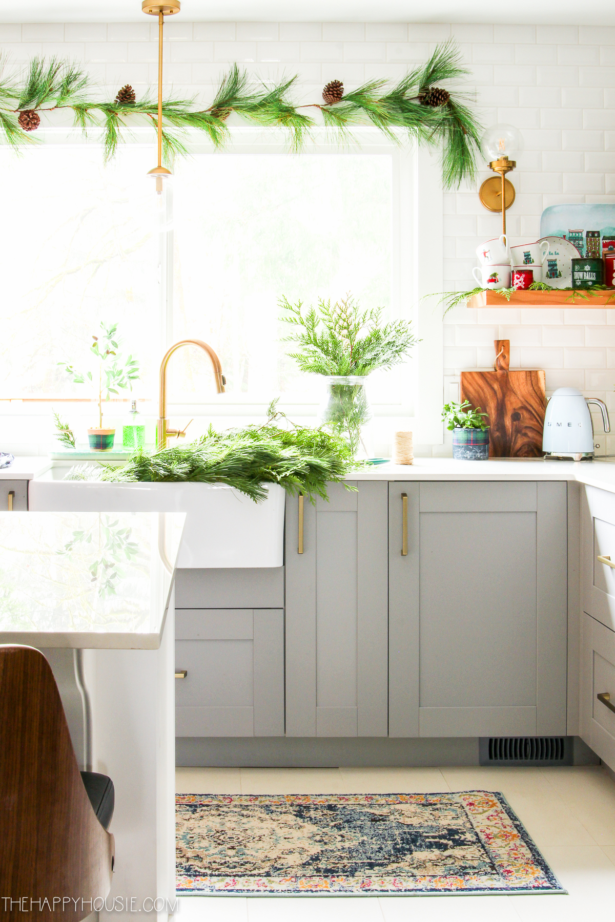There are gold pulls on the cabinets. A fresh greenery of garland is around the kitchen window.