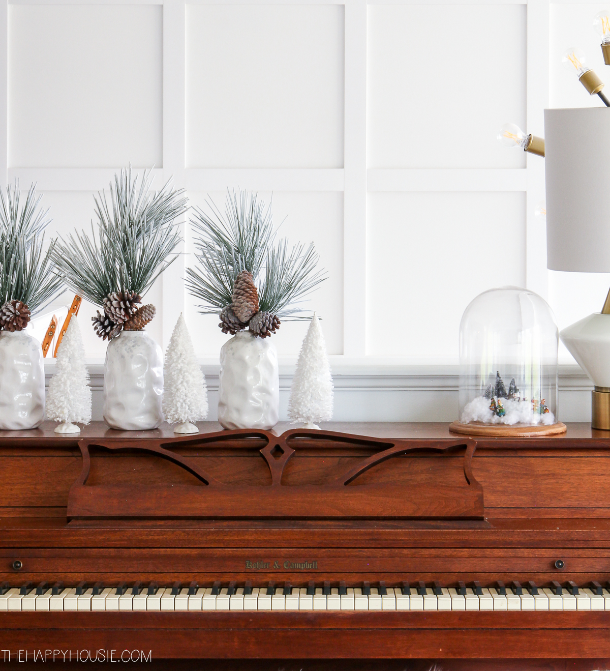 There are small white vases filled with flocked greenery and pine cones on the piano.
