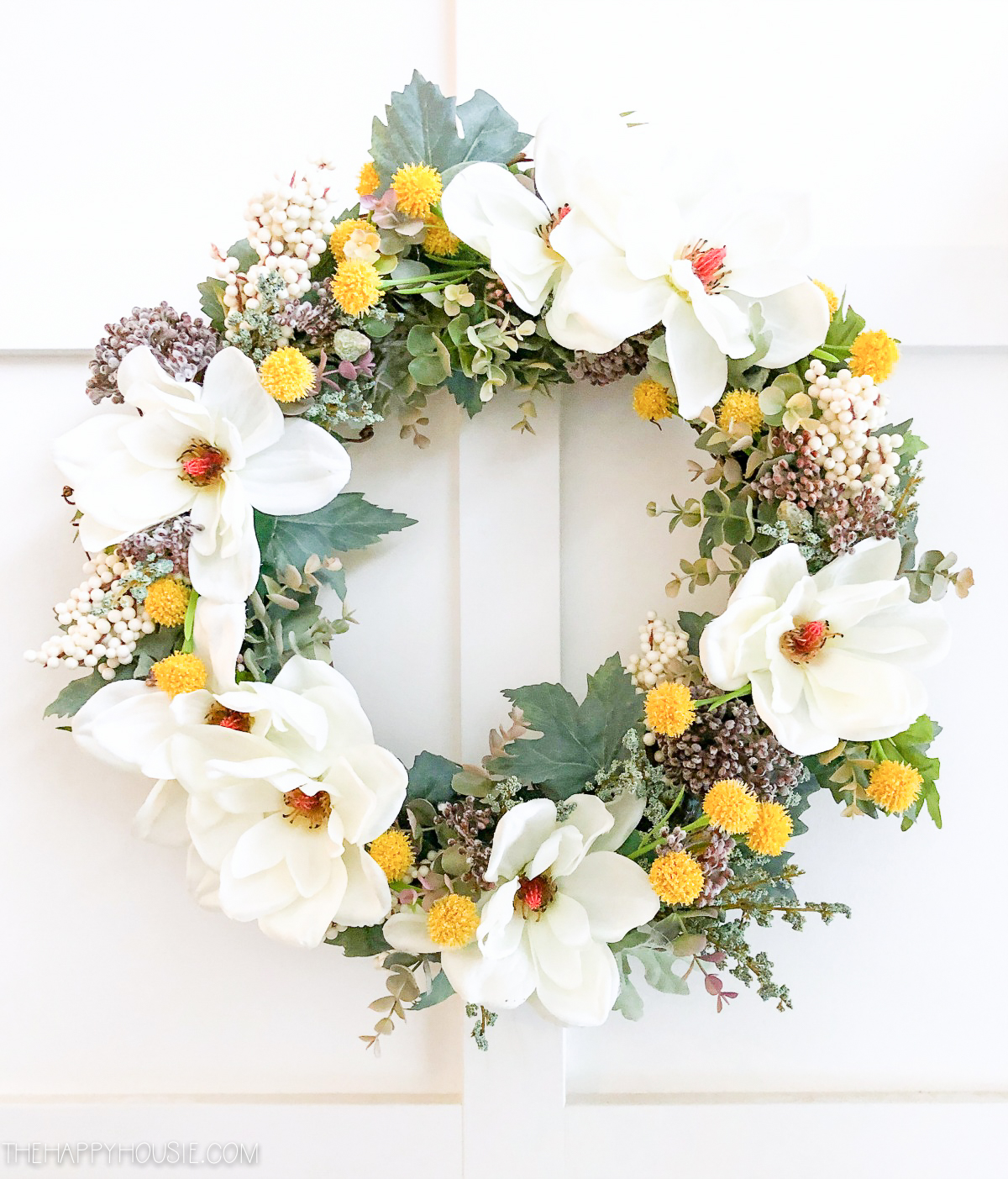 The wreath on a white door with the yellow flowers and greens.