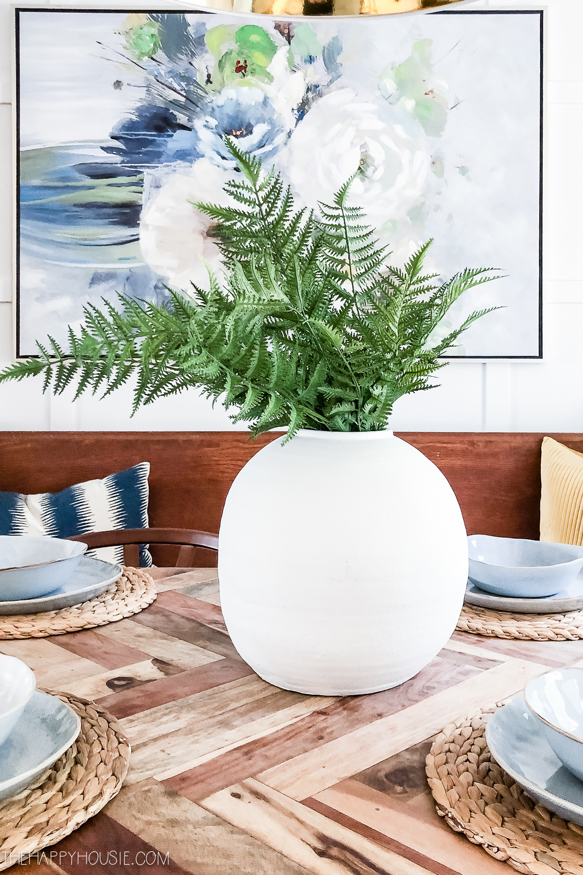 A white vase with ferns in it and a picture behind it on the table.