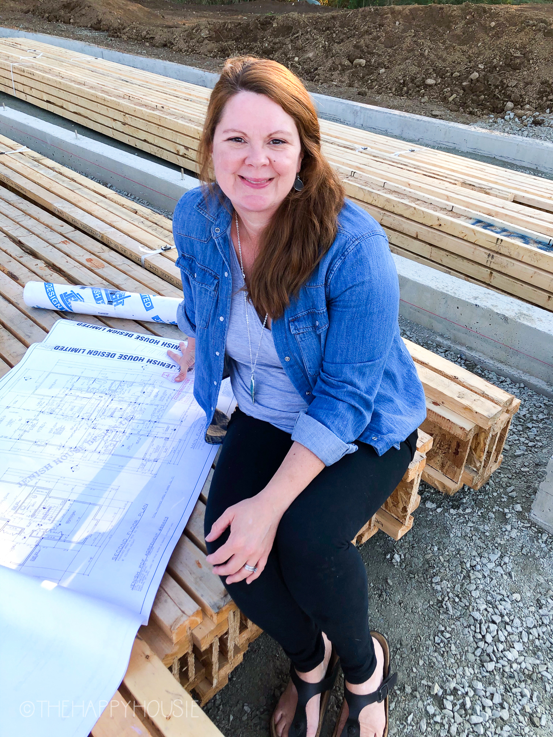 Krista sitting on a pile of wood with house plans laid out in front of her.
