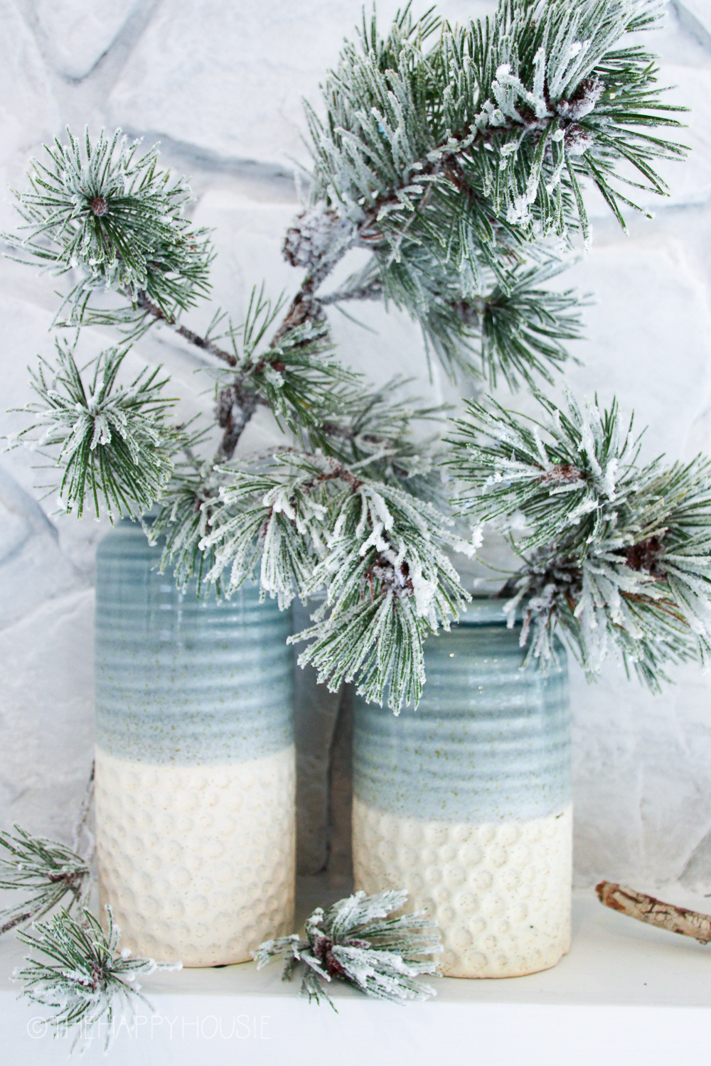 Two pottery vases with gradients of off white and green and blue with a flocked evergreen branch inside.