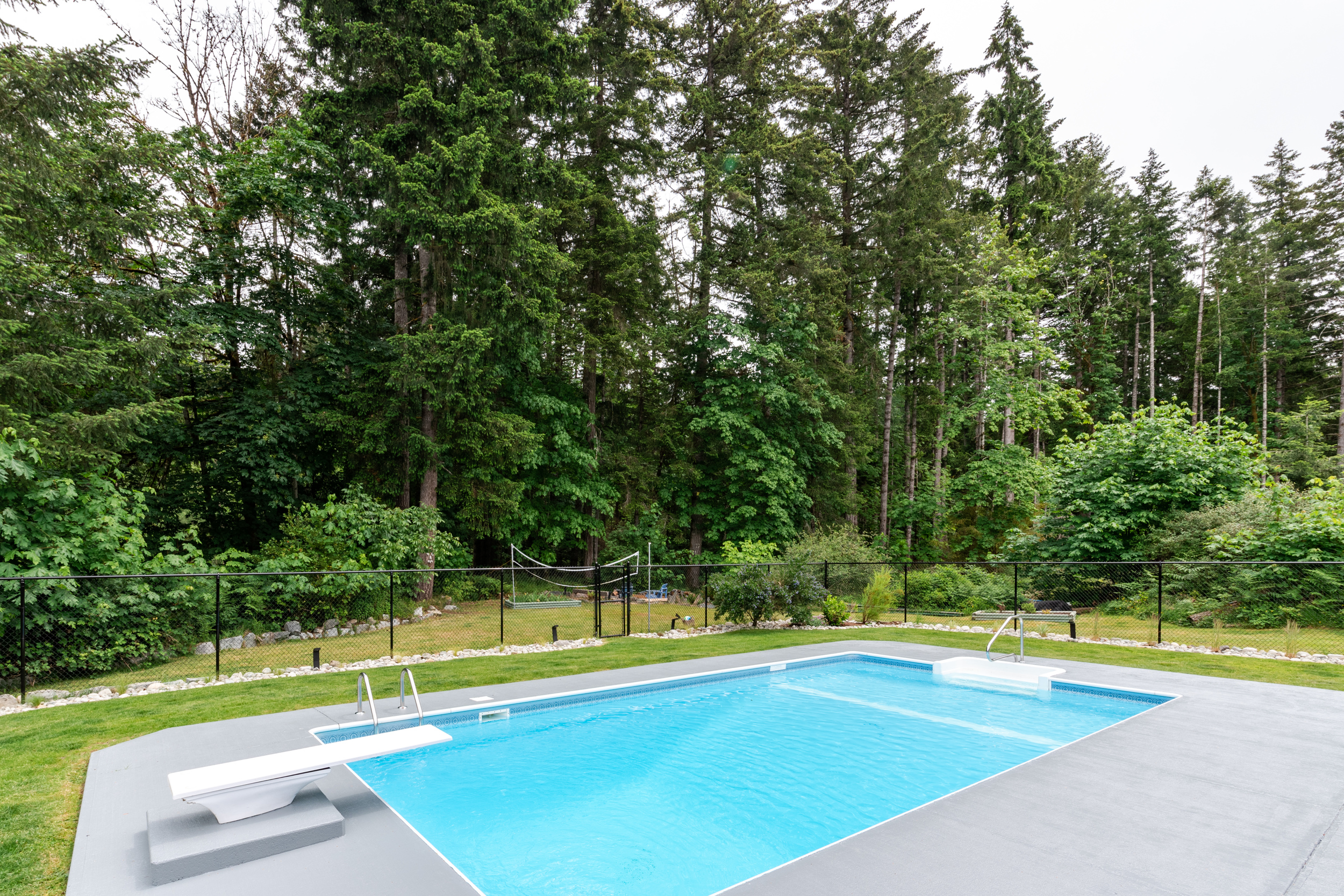 an in-ground swimming pool in a backyard with grey painted concrete pool deck and a forest in the backdrop