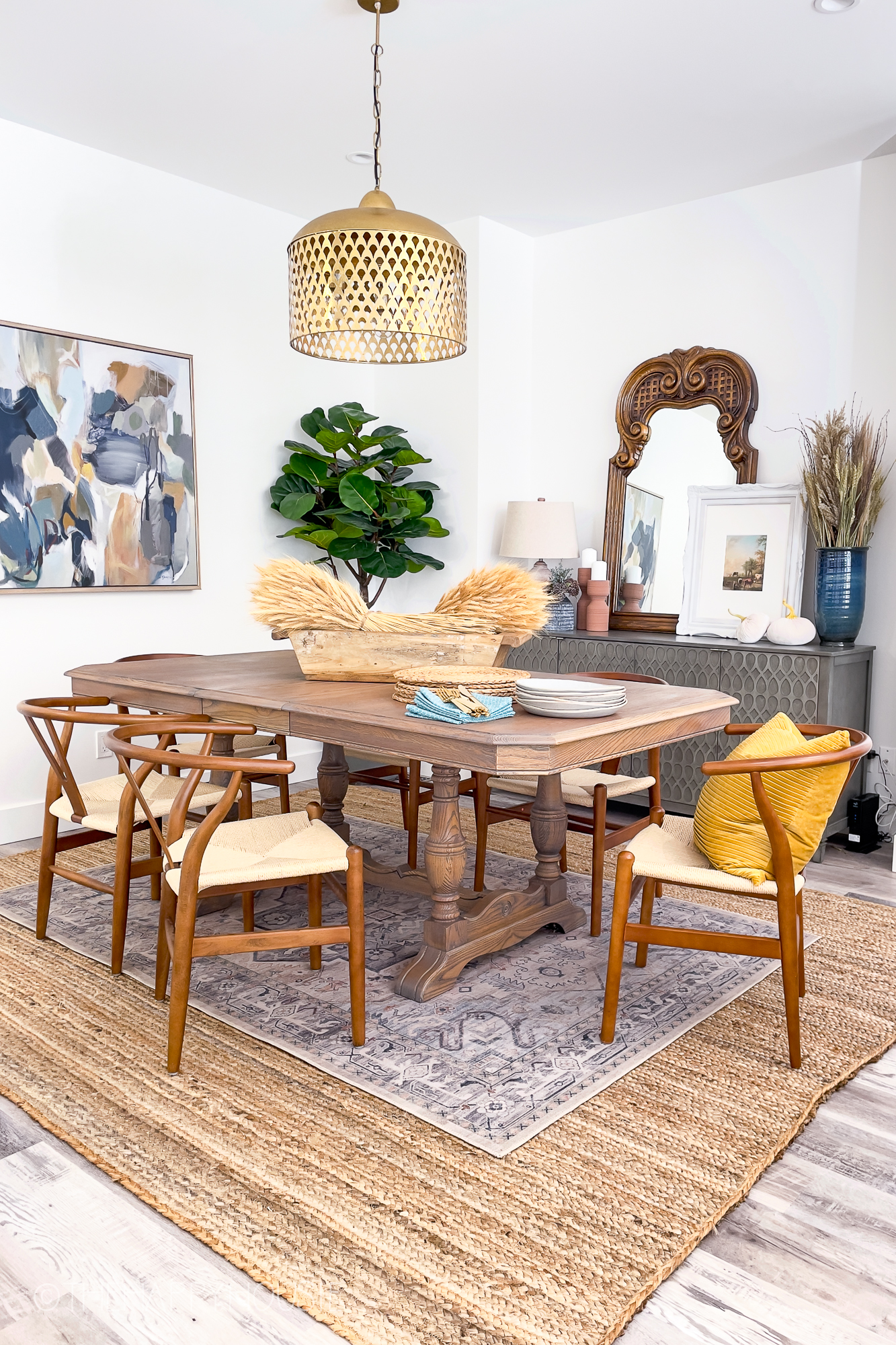 A wooden dining room table with a rug underneath and a gold light over the table.