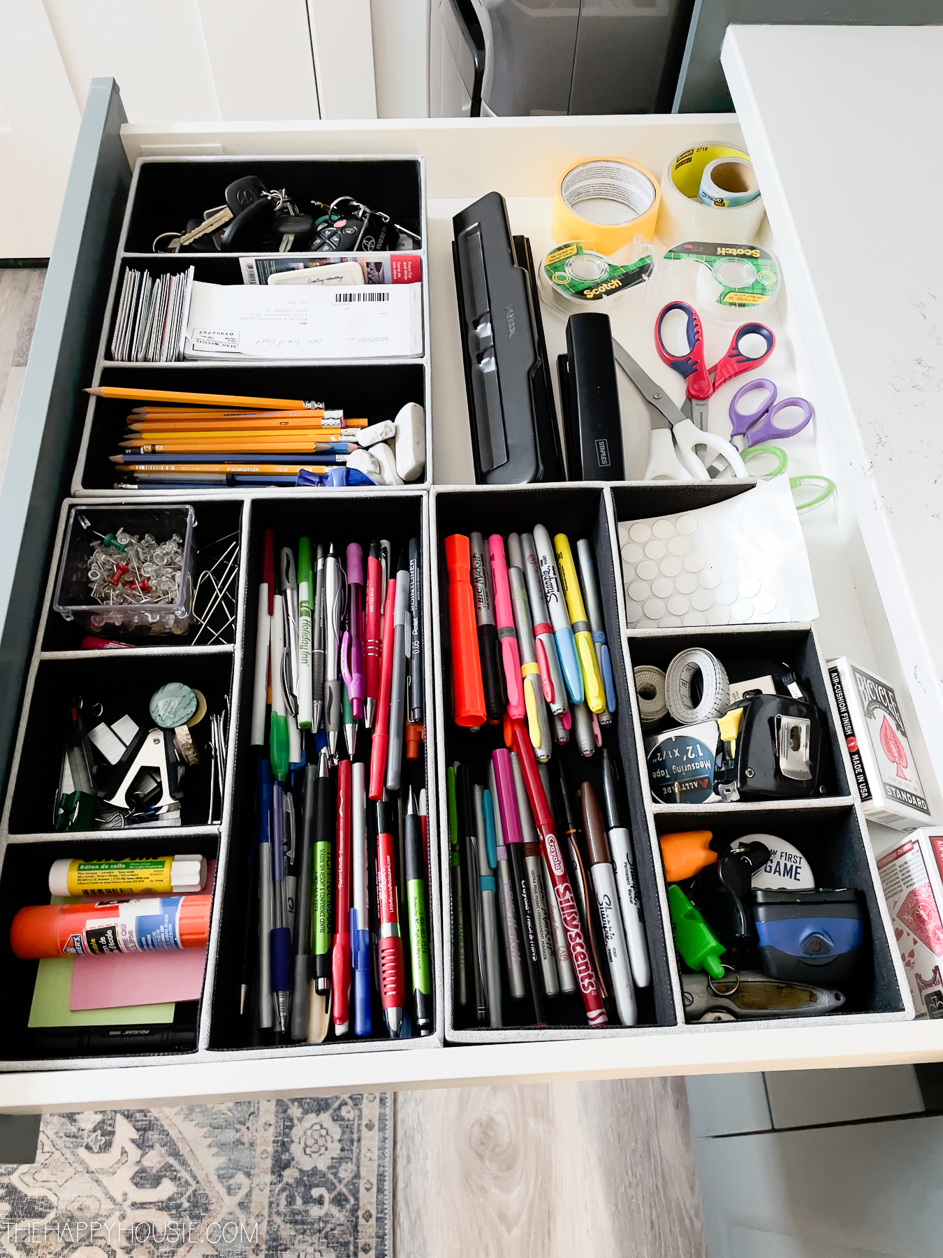 The office drawer open with pens, thumb tacks, scissors and tape.