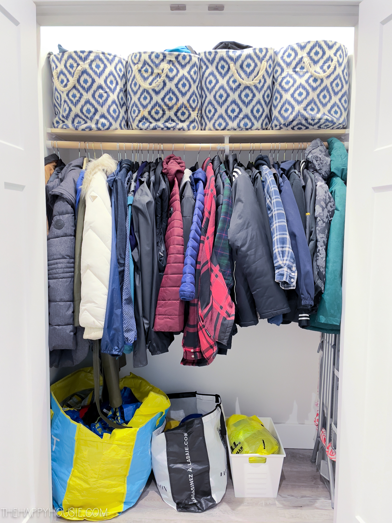 A closet by the door filled with coats, shopping bags and woven baskets on the top shelf.